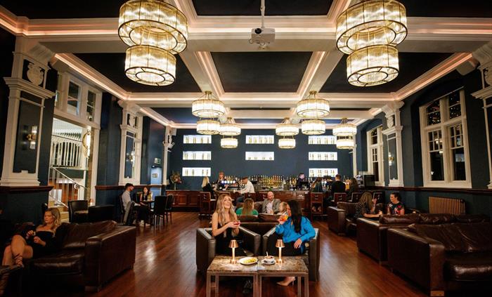 Image shows a large bar-lounge, with some guests sitting on sofas and drinking cocktails, whilst others sit at the bar at the rear.